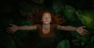 Девушка с рыжими волосами и веснушками стоит среди зелёных листьев, раскинув руки и закрыв глаза, выражая чувство свободы и гармонии с природой.