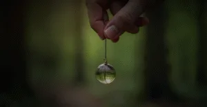Рука держит прозрачный стеклянный шарик на нитке на фоне леса, создавая атмосферу загадочности и волшебства.