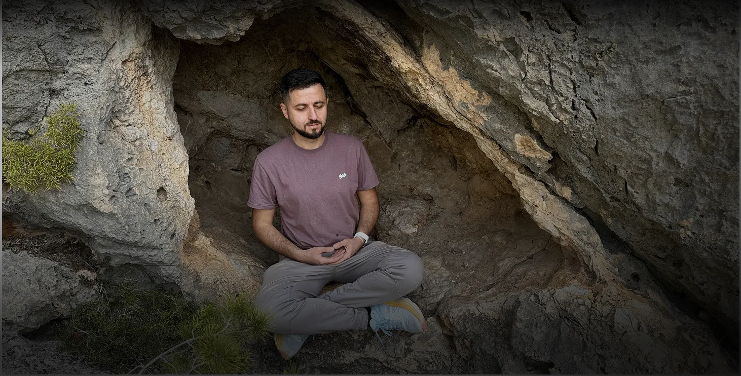 Мужчина медитирует в природной пещере, окружённой камнями и растительностью, что создает атмосферу спокойствия и гармонии с природой.