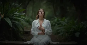 Женщина в белом одеянии медитирует на скамейке в лесу среди зелёных растений, создавая атмосферу спокойствия и гармонии.