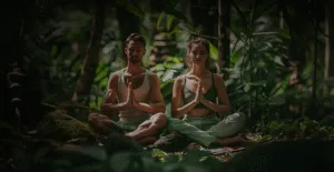 Молодая пара медитирует в тропическом лесу, окруженная зелеными растениями, что создает атмосферу спокойствия и гармонии.